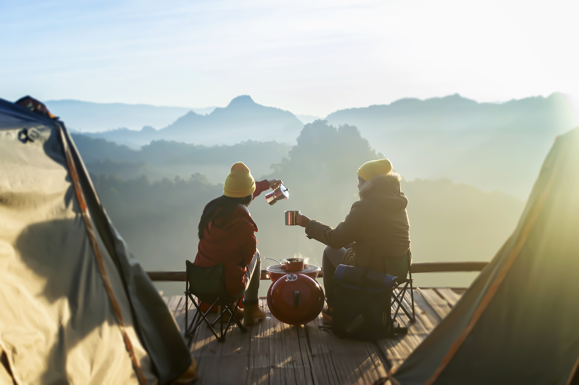 Two people sit outside their tents on a wooden deck with a mountainous landscape in the background. They toast with mugs while wearing winter clothes, and a small red camping stove sits between them. The scene is bathed in soft morning light.