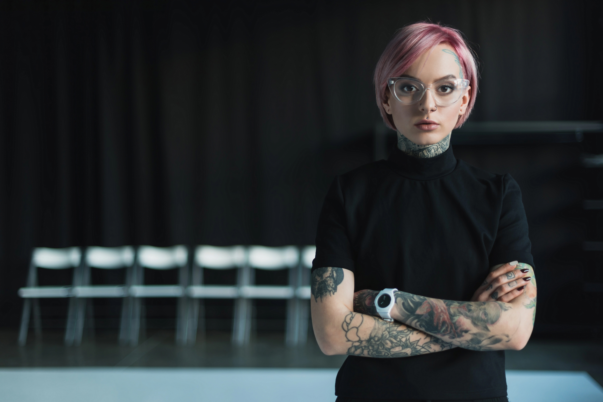 A person with short pink hair, wearing glasses and a black turtleneck, stands with arms crossed. They have multiple tattoos on their arms and neck. The background is dim, with white chairs aligned in a row.