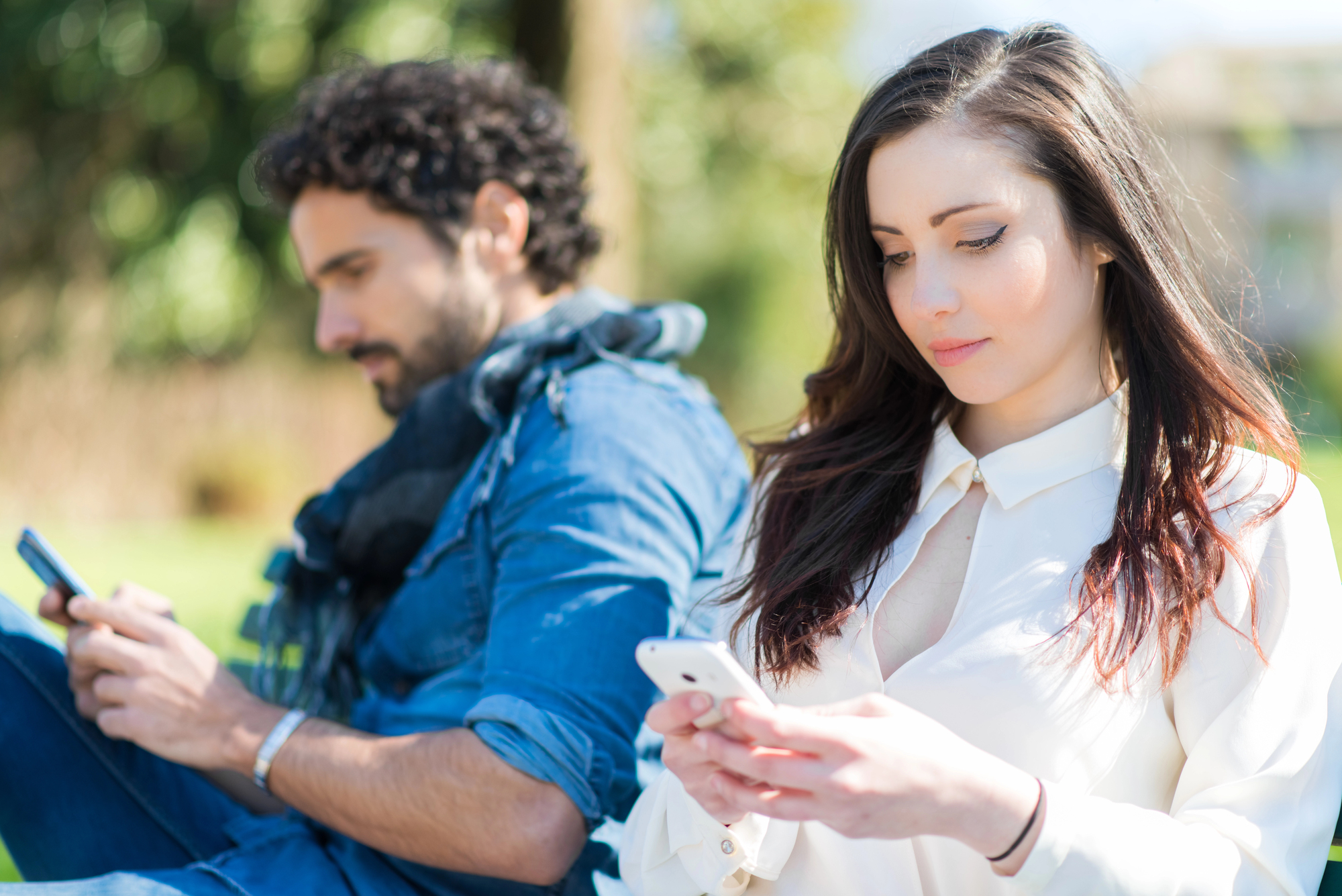 A woman and a man sit back-to-back outdoors on a sunny day, both engrossed in their smartphones. The woman, with long dark hair, wears a white blouse. The man, with curly dark hair and a beard, is wearing a denim shirt with a scarf around his neck.