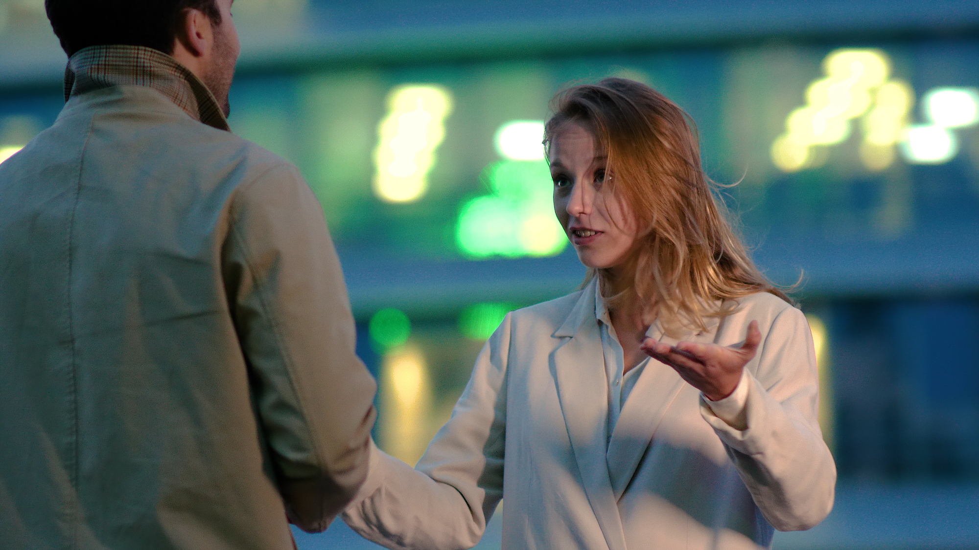 A woman with blonde hair, wearing a light-colored coat, gestures with her right hand and appears to be talking to a man in a beige jacket. The conversation takes place outdoors, with blurred lights illuminating the background.