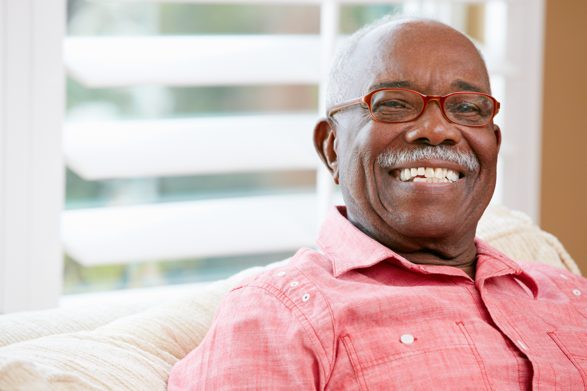 A smiling elderly man with short gray hair and a mustache is sitting on a couch. He is wearing a pink button-up shirt and red-framed glasses. The background features white blinds and a softly focused interior setting.