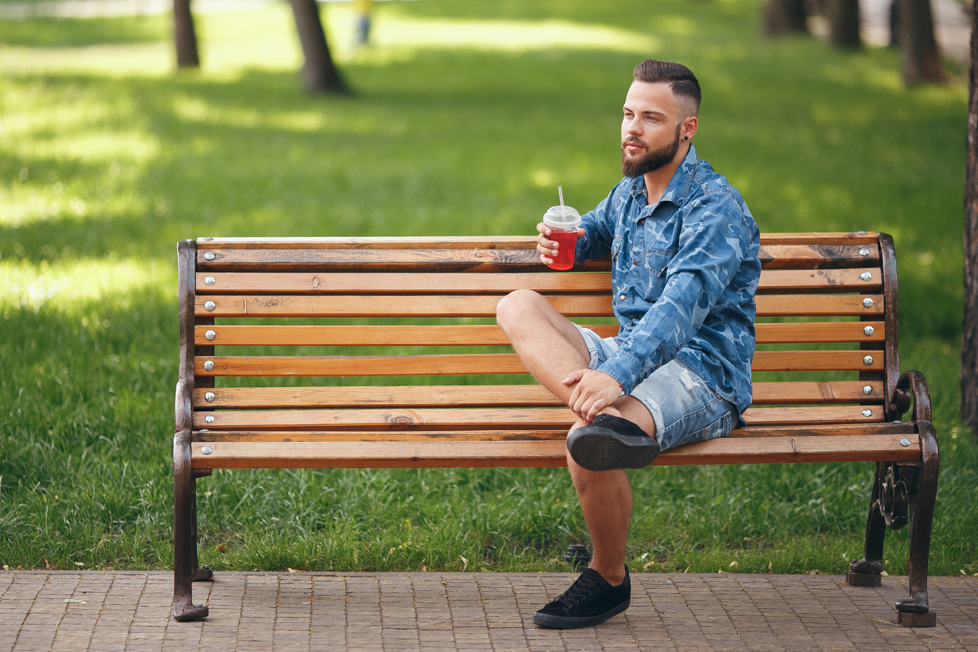 A man with a beard sits on a wooden park bench, holding a cold beverage with a straw, wearing a blue denim shirt and shorts, and black shoes. He's gazing into the distance on a sunny day with lush green grass and trees in the background.