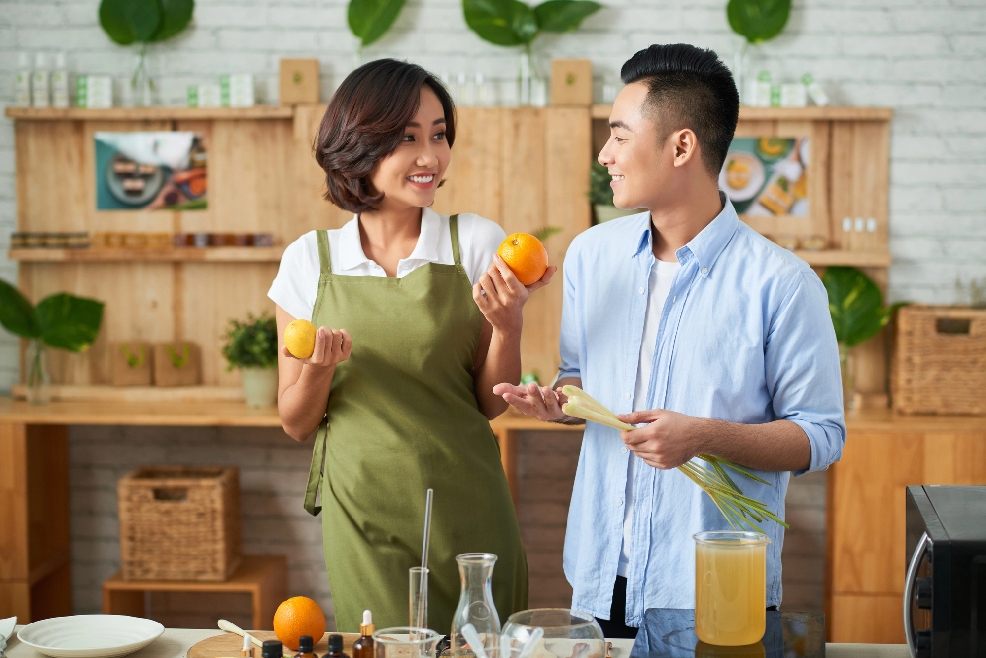 A woman in a green apron and a man in a light blue shirt stand in a kitchen, smiling at each other. The woman is holding two oranges, and the man is holding a bunch of lemongrass. The kitchen has wooden shelves, green plants, and various kitchen utensils.