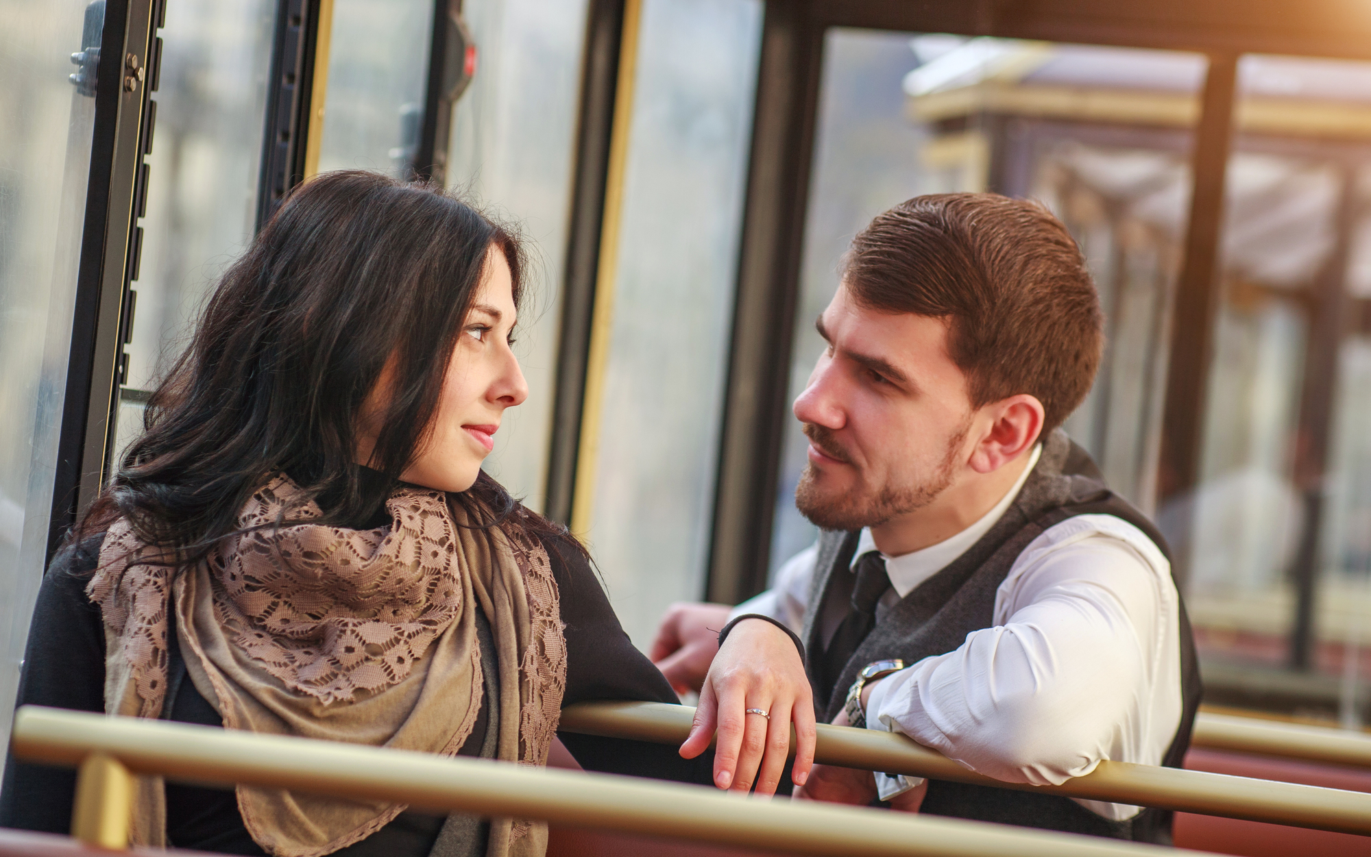 A woman with long dark hair and a scarf gazes at a man with short brown hair and a beard. They are sitting closely in a bus or tram, leaning on a handrail, looking at each other with slight smiles. The sunlit window suggests it's daytime.