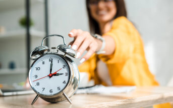 A person in a yellow jacket reaches out to turn off a silver analog alarm clock on a wooden table. The clock shows the time as 7:12. The background is slightly blurred, indicating focus on the clock.