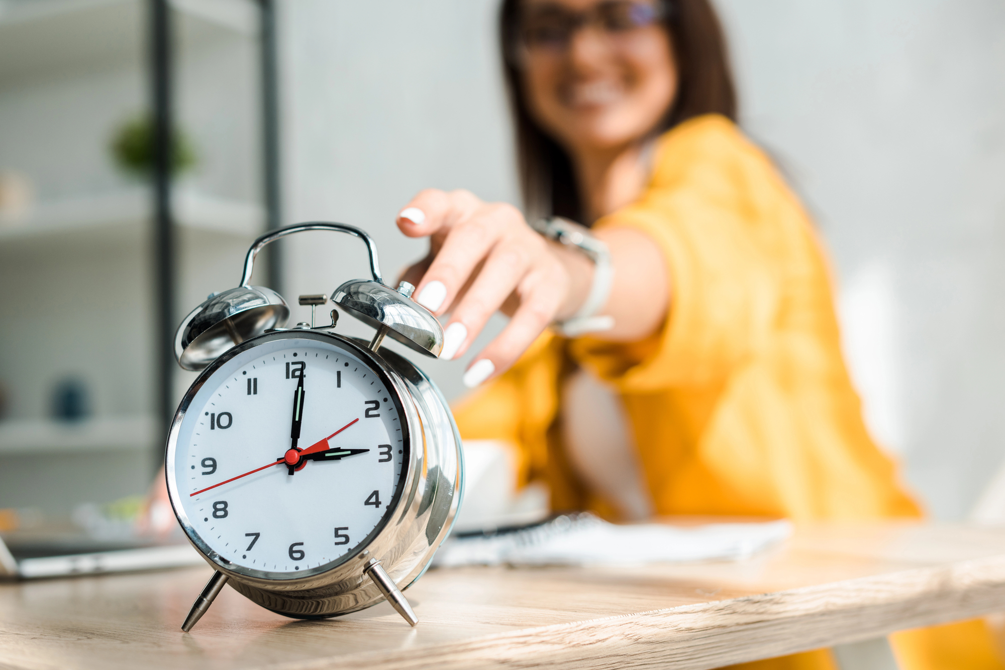 A person in a yellow jacket reaches out to turn off a silver analog alarm clock on a wooden table. The clock shows the time as 7:12. The background is slightly blurred, indicating focus on the clock.