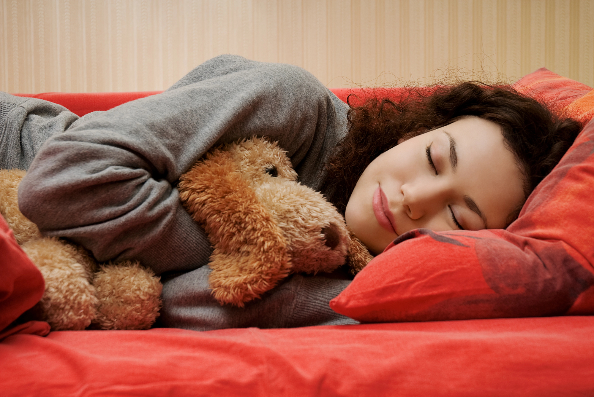 A person with curly hair is lying on a red bed, hugging a brown stuffed animal. They are wearing a grey sweatshirt and appear to be peacefully sleeping. The background features a light-colored, vertically striped wallpaper.