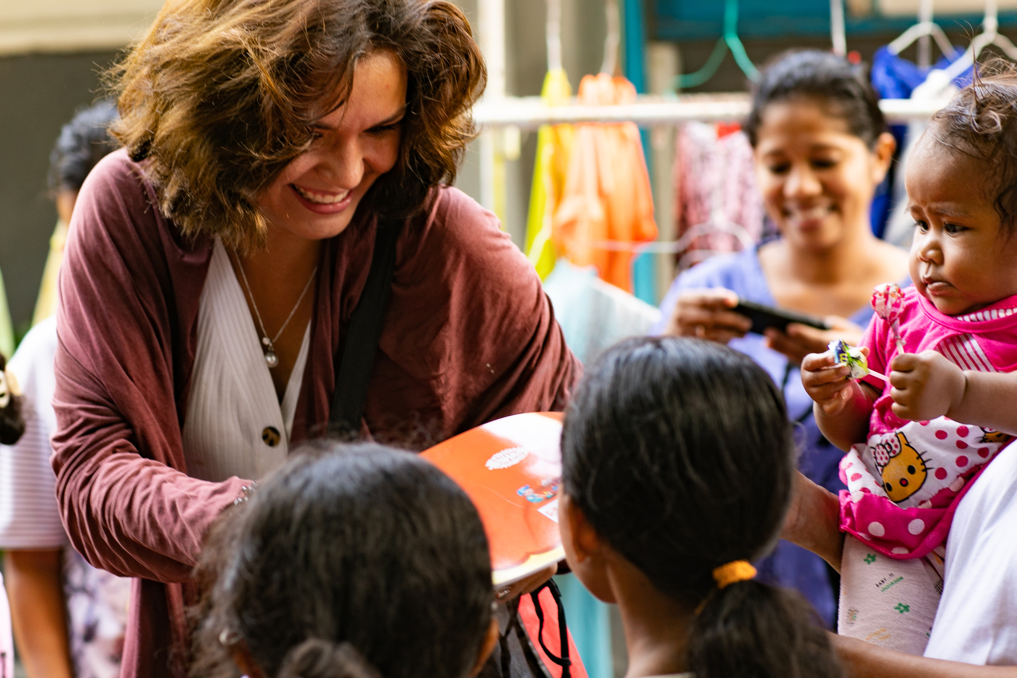 A woman with curly hair and a pink sweater smiles as she hands a book to a group of children. The children gather around her eagerly, with one holding a baby dressed in pink. Brightly colored clothes hang in the background, adding a cheerful atmosphere.