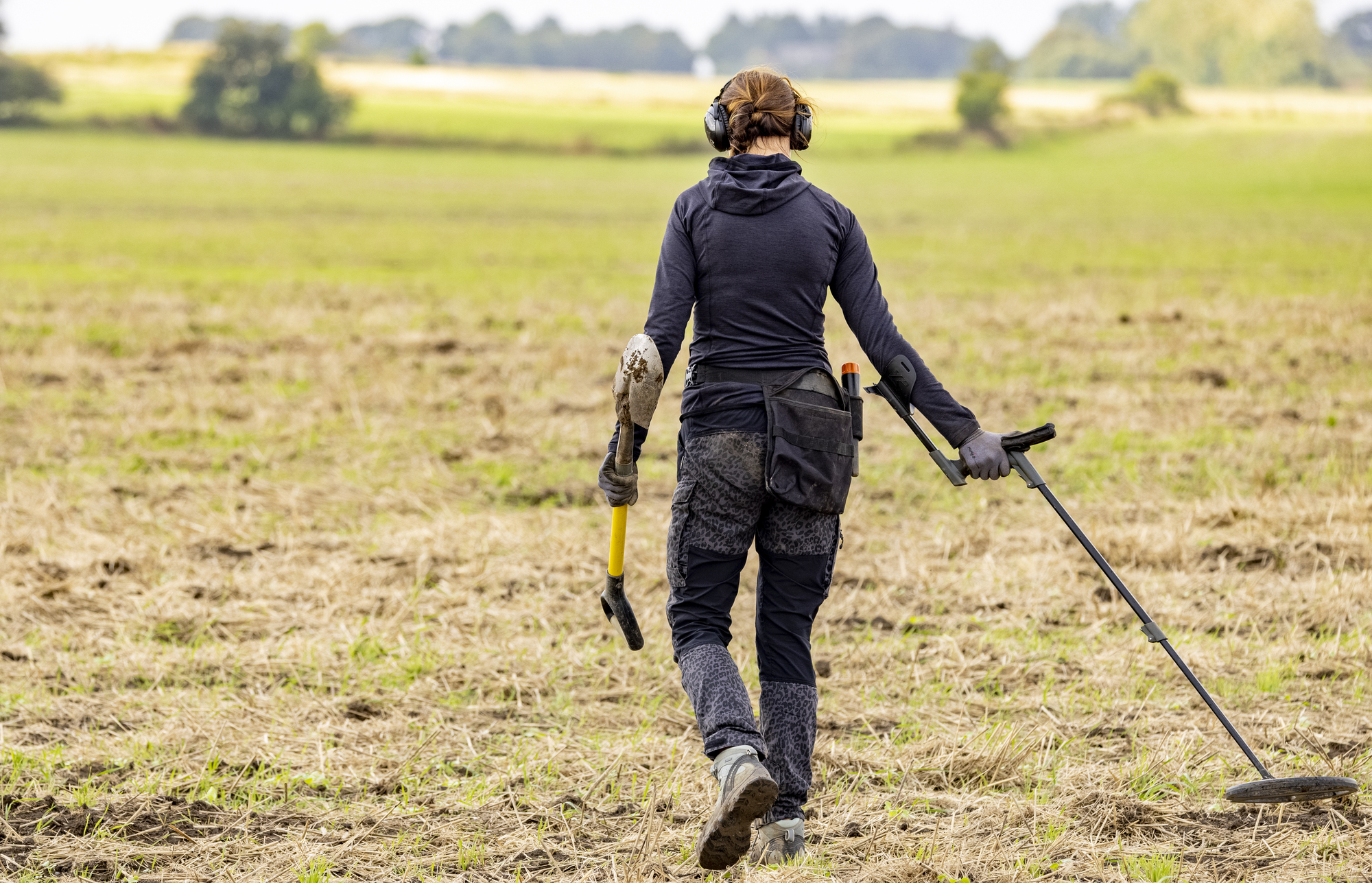 A person dressed in dark clothing, wearing headphones, and holding a metal detector is walking across a large, open, grassy field. They also carry a small digging tool in their other hand. The background shows distant trees and an overcast sky.