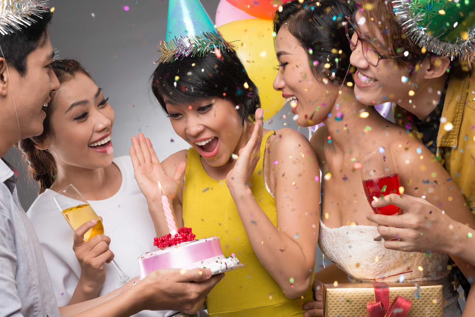 A group of five people celebrate a birthday, three women and two men. One woman in the center, wearing a party hat, is excitedly looking at a small pink cake with a candle. Confetti is falling, and festive balloons are in the background.