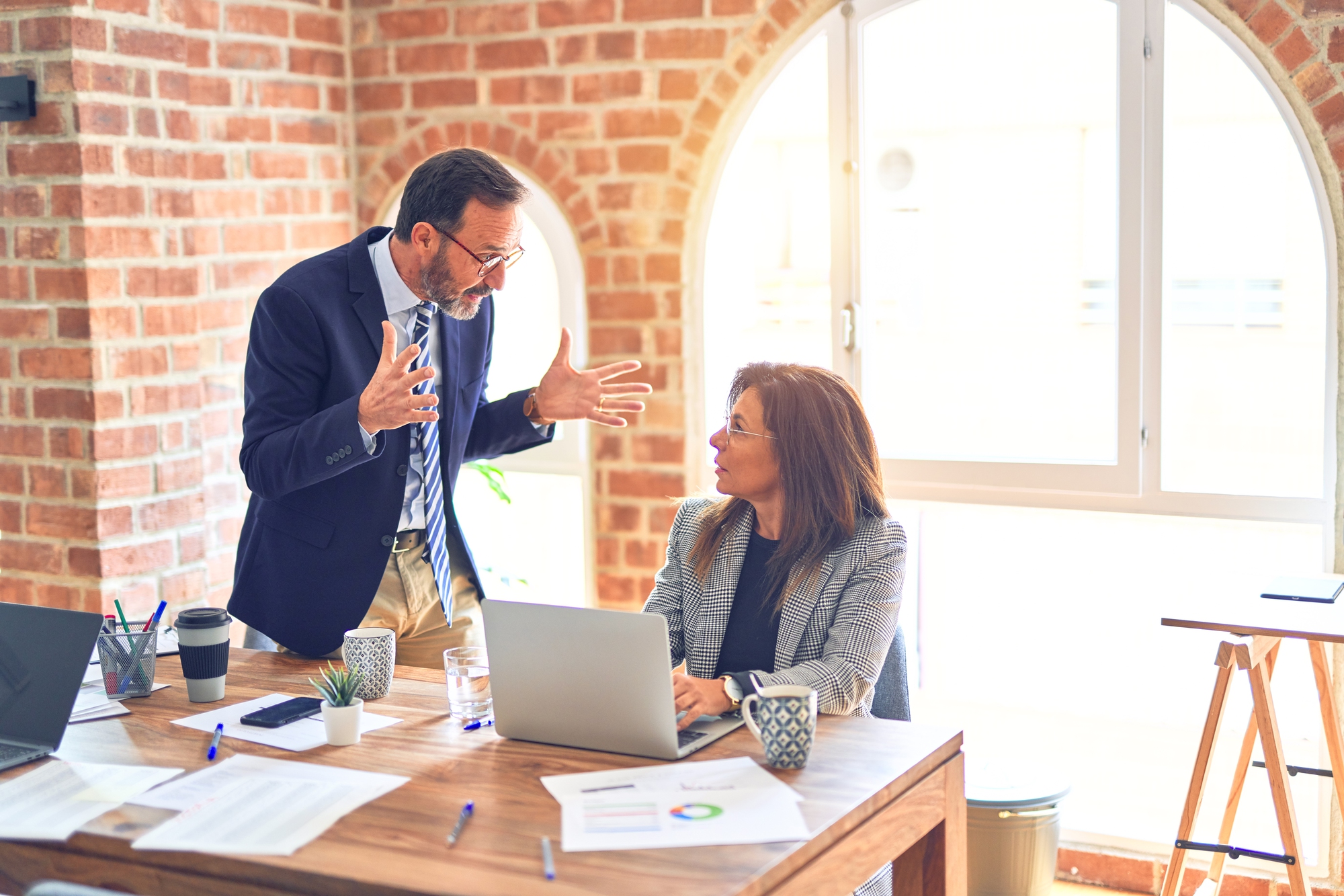 A man in a suit gestures passionately while talking to a woman seated at a desk with a laptop. Both are in a brightly lit room with brick walls and large windows. The desk is cluttered with papers, pens, a coffee cup, and other office items.