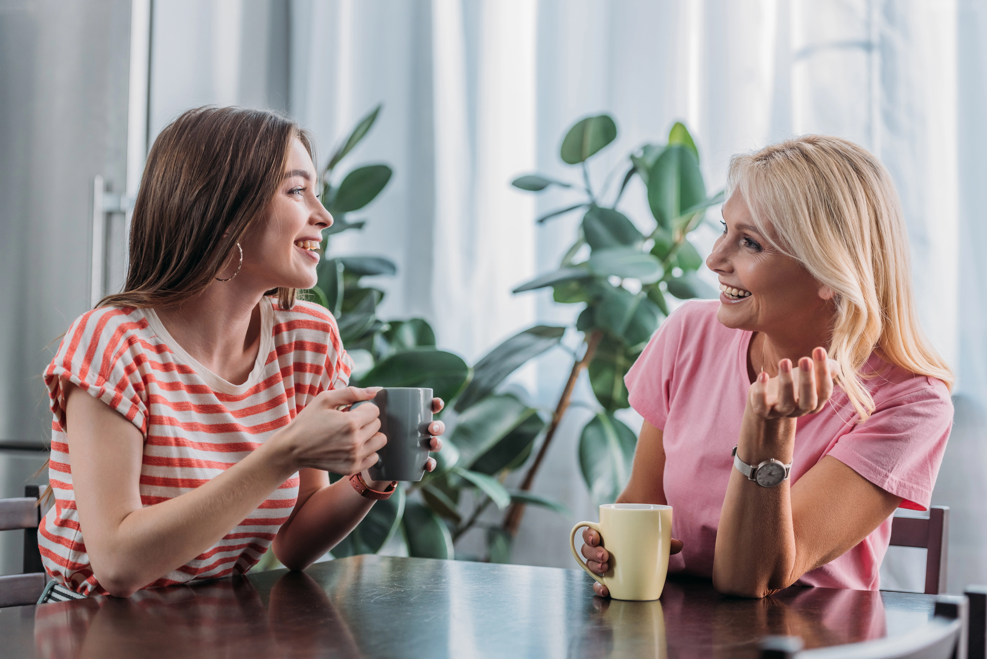 Two women sitting at a table holding coffee mugs and engaged in conversation. One wears a red and white striped shirt while the other wears a pink shirt. There are plants and curtains in the background, and both women appear happy and engaged.