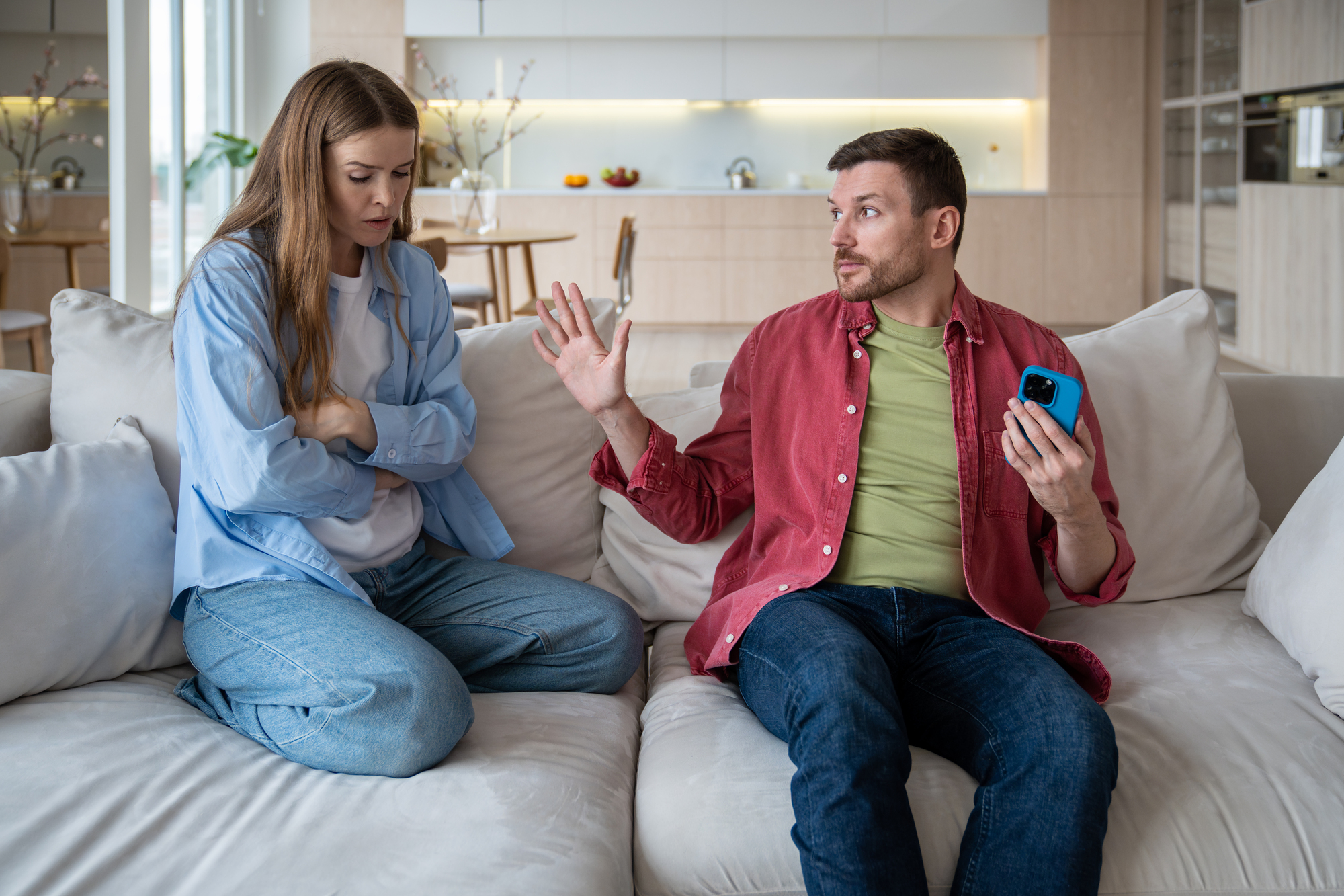 A woman with folded arms looks upset while sitting on a couch next to a man who is gesturing with his hand and holding a smartphone. They are in a modern living room with a kitchen in the background.