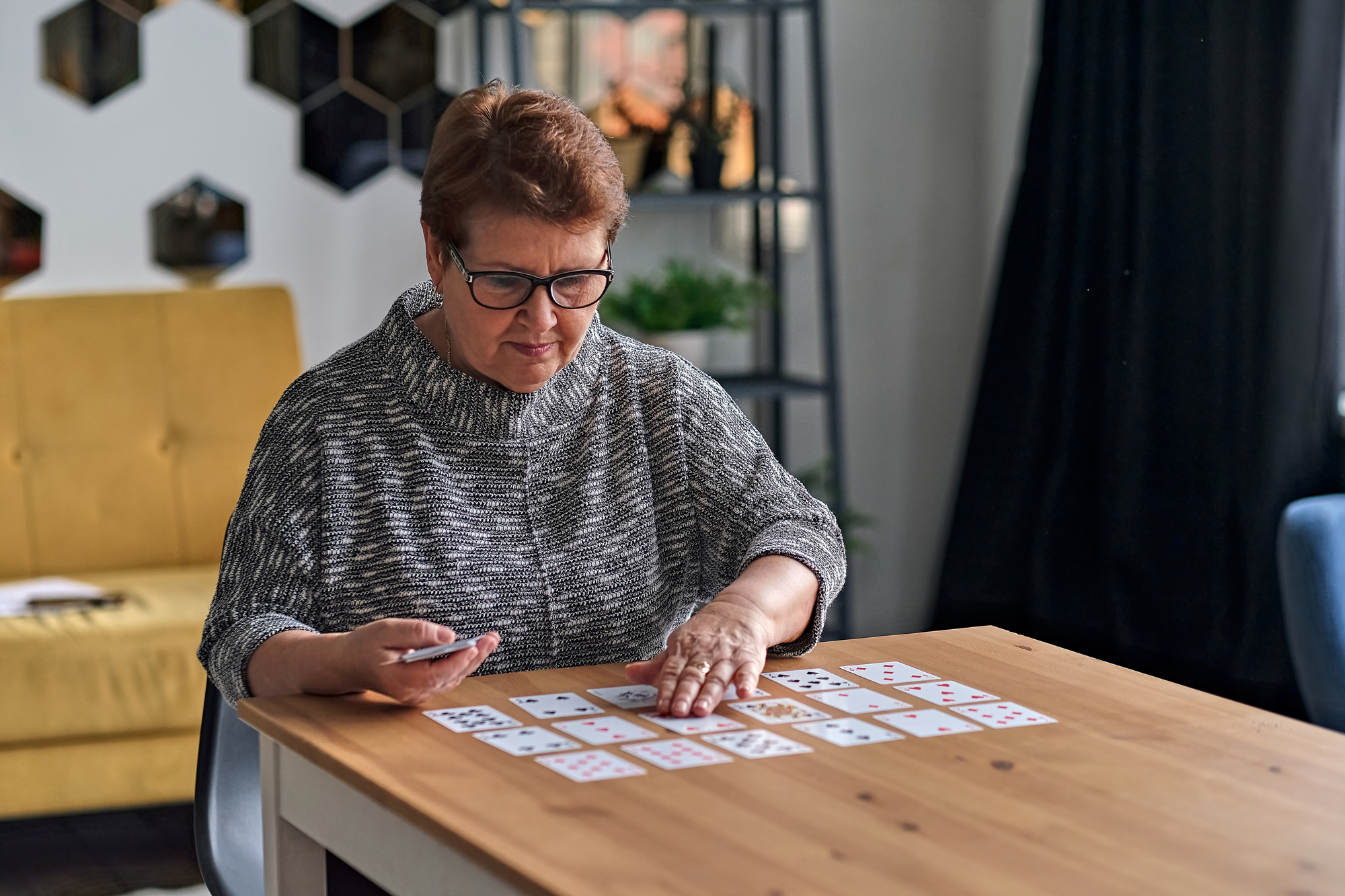 A woman with short hair and glasses sits at a wooden table, concentrating on arranging playing cards in a solitaire game. She wears a gray sweater and is in a cozy room with a yellow chair and modern decor in the background.