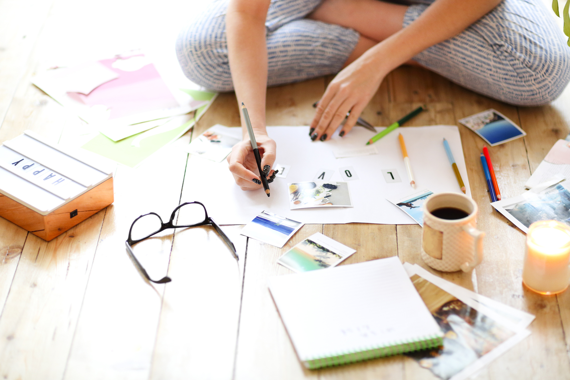 A person sits cross-legged on the floor, surrounded by papers, photos, and stationery. They are writing on a piece of paper, with a coffee mug, eyeglasses, and a candle nearby. The scene appears to be a creative workspace.