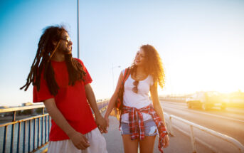 A man and woman are holding hands while walking on a bridge during sunset. The man, wearing a red T-shirt and beige shorts, has long dreadlocks. The woman, dressed in a white tank top, plaid shirt tied at the waist, and denim shorts, is smiling at him. A car is visible in the background.