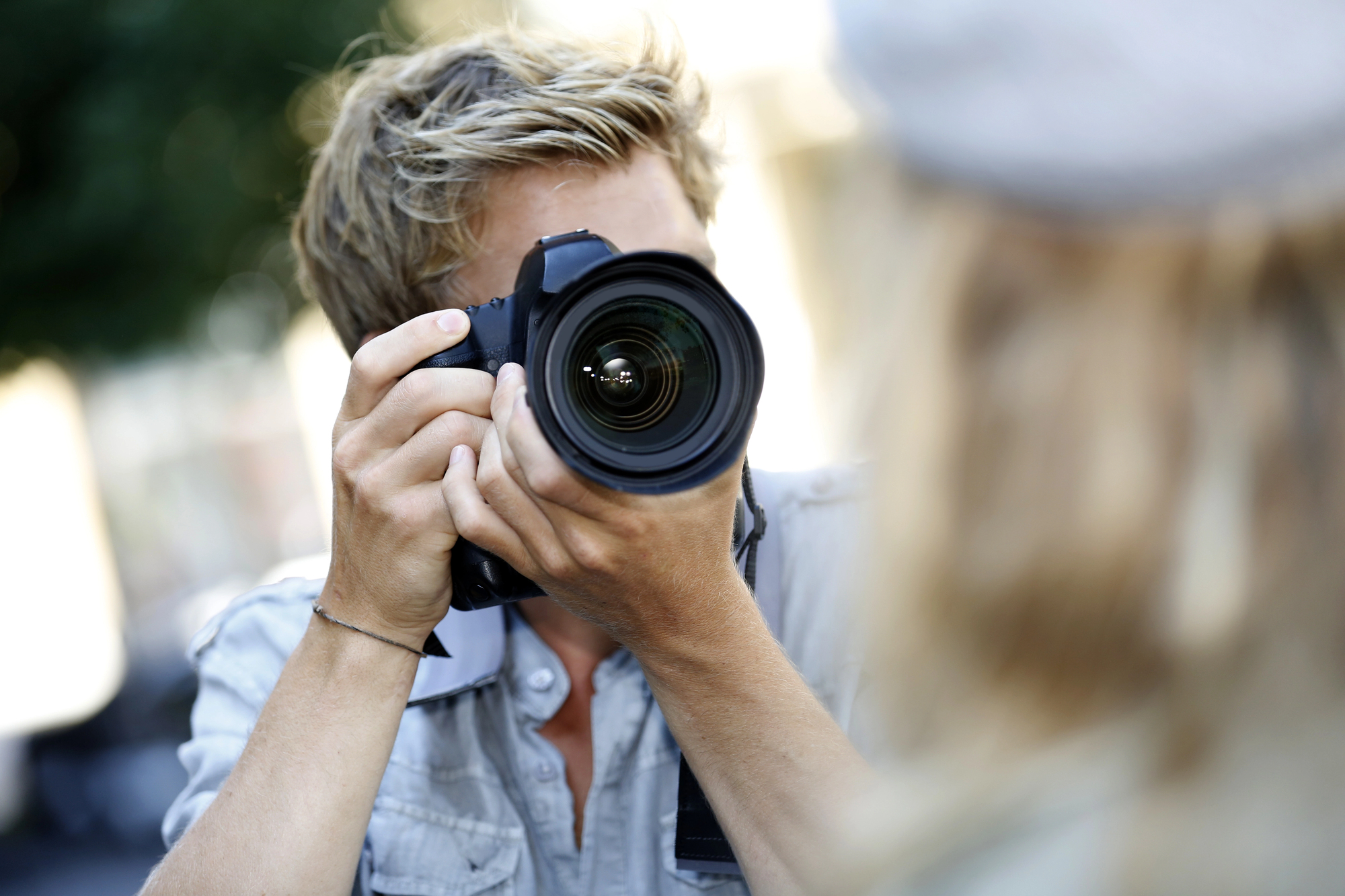 A person with light, tousled hair holds a camera up to their face, aiming the lens directly at the viewer. The individual is outdoors and wears a light-colored shirt, with some greenery blurred in the background. The focus is on the camera and the person's hands.