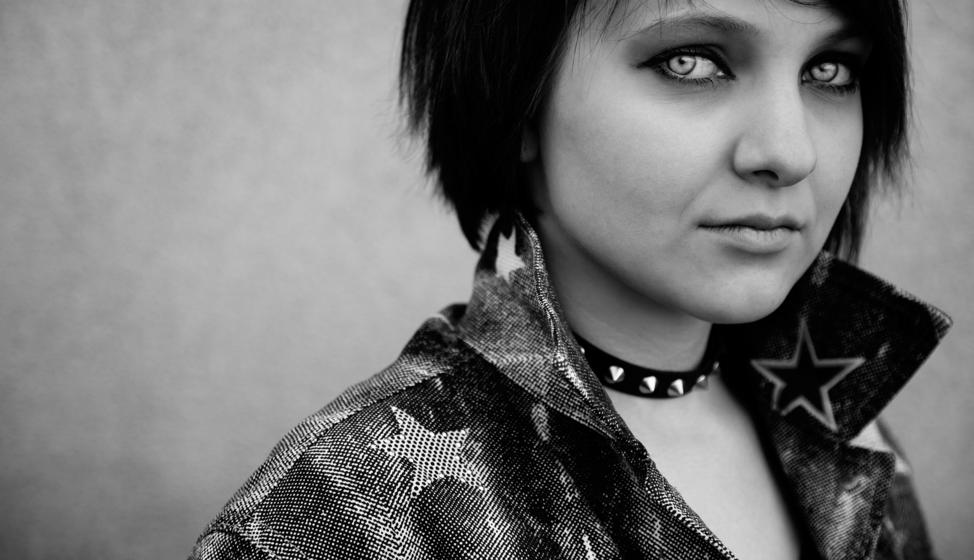 Black and white photo of a person with short dark hair, wearing a spiked choker necklace and a denim jacket with star patterns. The image is zoomed in, highlighting the person's face and the details of their jacket. The person gazes slightly off-camera.