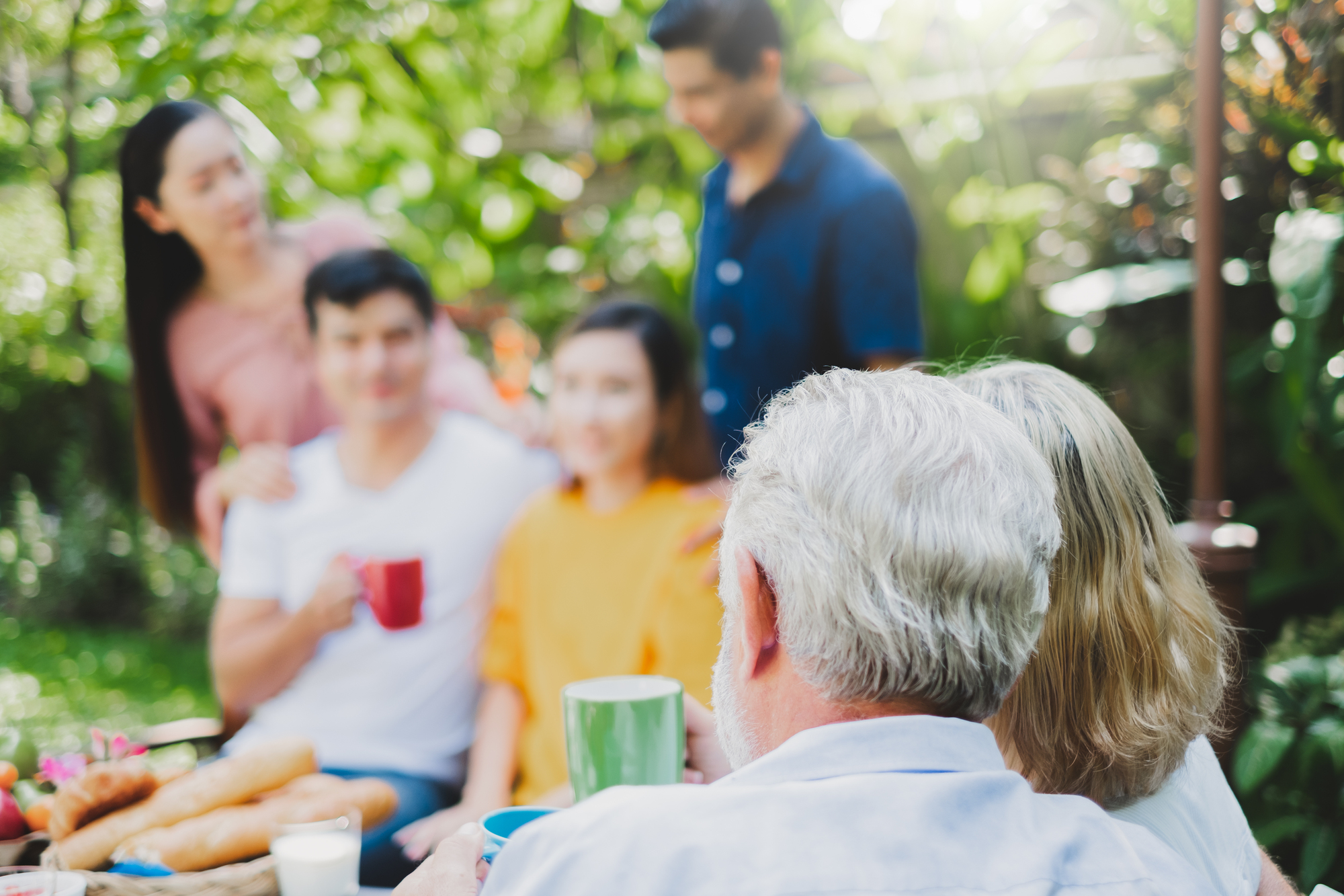 A group of six people enjoys a backyard gathering. In the foreground, an older couple is seated and holding coffee mugs. In the background, four younger individuals are standing and smiling, with greenery surrounding them. The scene is relaxed and cheerful.