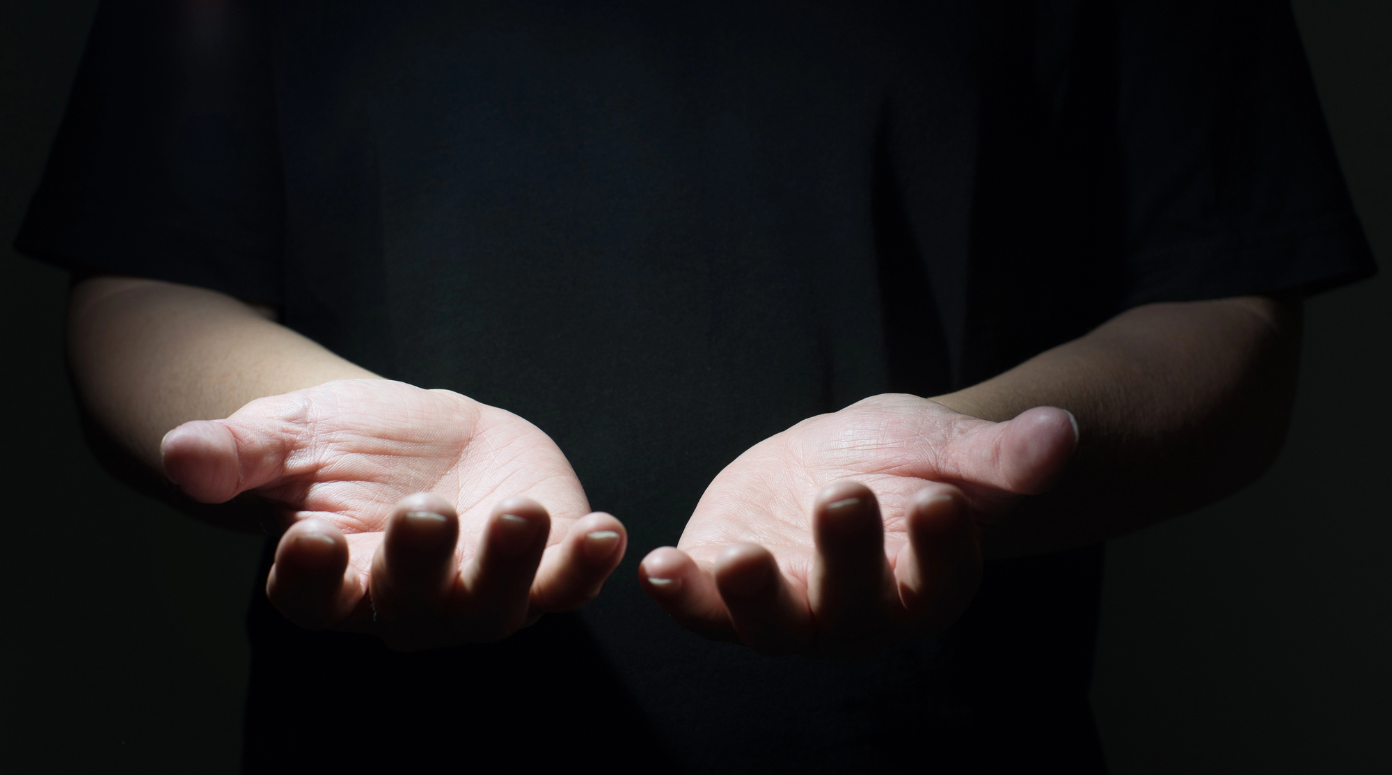 A close-up of a person wearing a dark shirt with their hands outstretched and palms open, illuminated against a dark background. The image focuses on the hands, highlighting the details and texture of the skin.