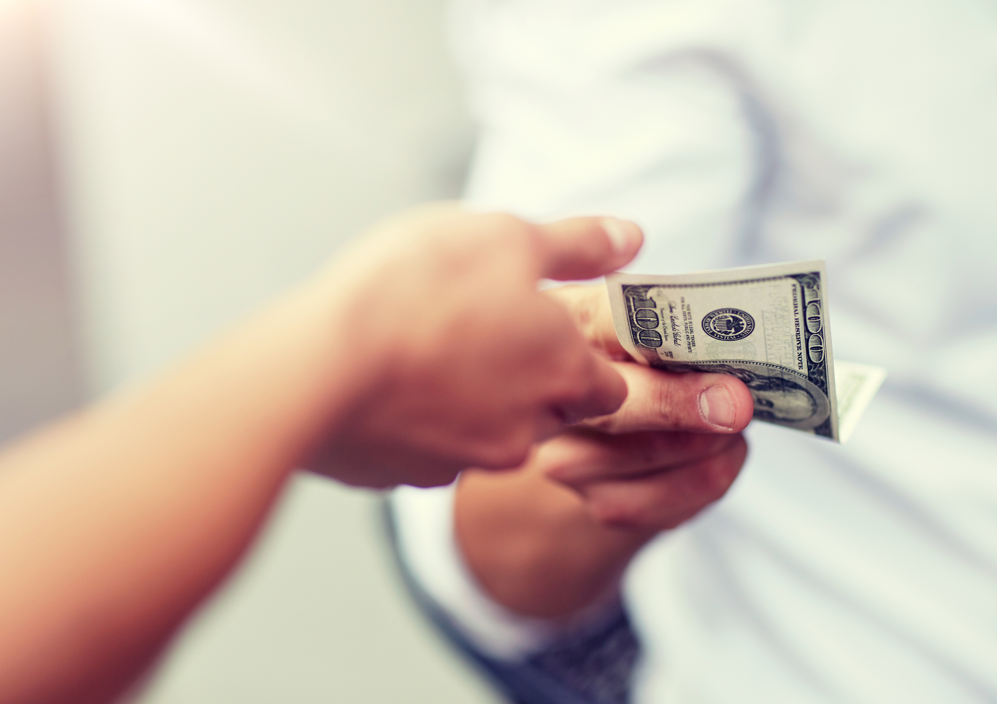 Close-up of one person handing a $100 bill to another person. The money is being exchanged against a light, blurred background. The person handing the money has a thumb and forefinger visible, while the recipient's hand is partially shown.