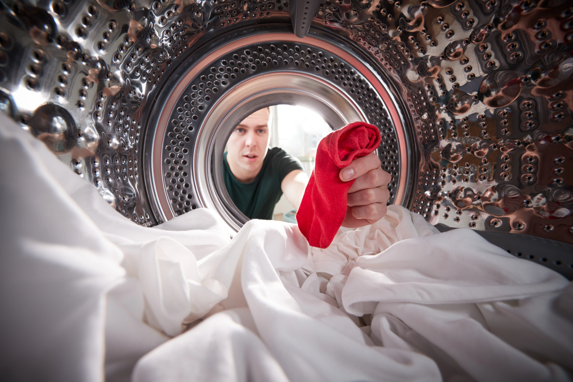 View from inside a washing machine drum as a person reaches in to remove a single red sock among a pile of white laundry. The person appears to be surprised or concerned. The drum's metallic interior and the door's circular frame are visible.