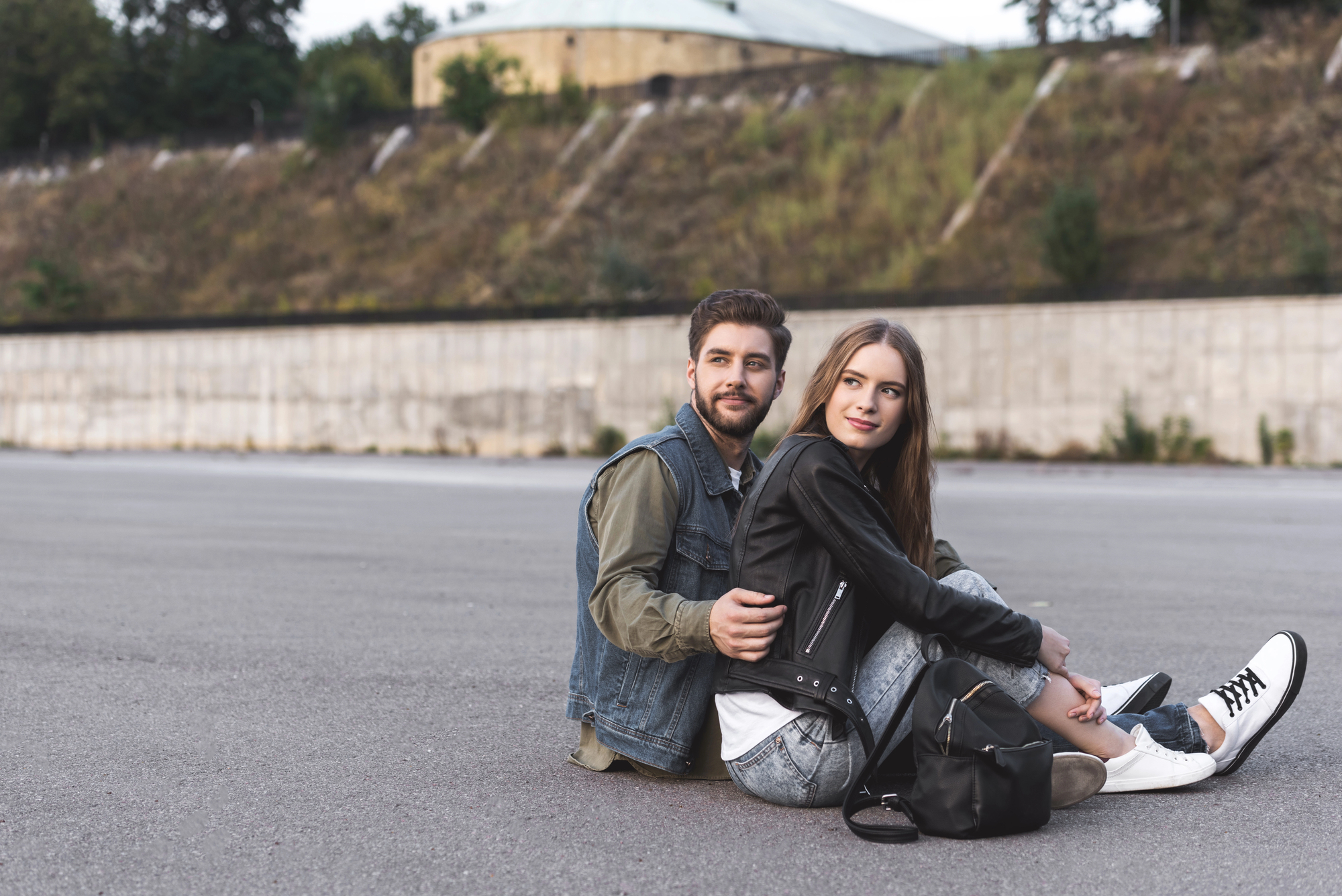 A young couple sits on an empty paved surface outdoors, leaning against each other. The man, with a beard, wears a denim jacket over an olive shirt. The woman, with long hair, wears a black leather jacket and white sneakers. They appear content and relaxed.