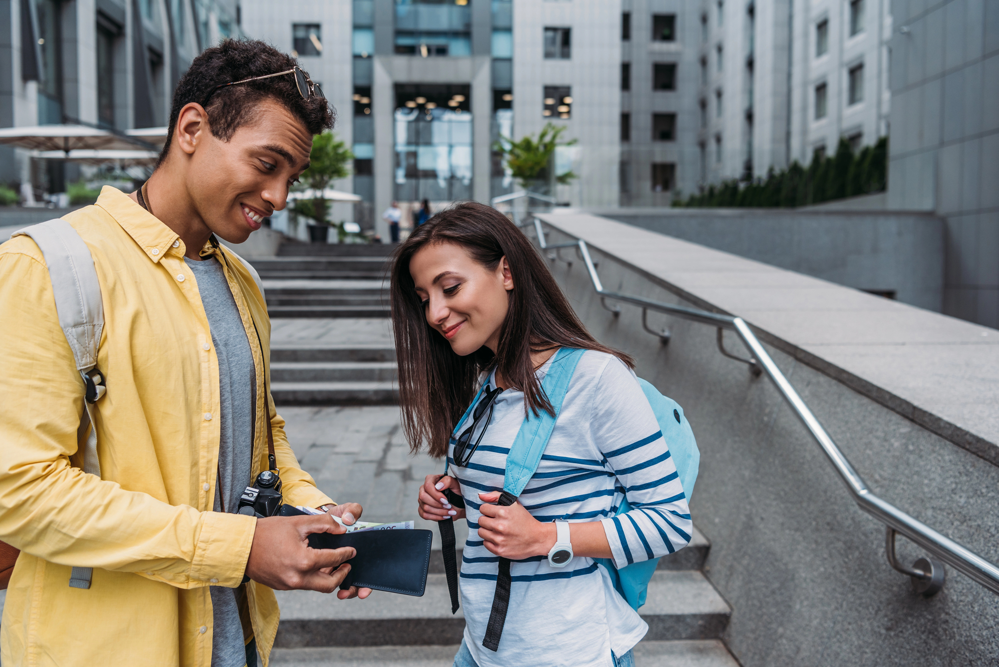 A young man and woman stand outdoors in an urban setting, smiling as the man shows something on his phone to the woman. They both wear casual attire with backpacks, appearing to be students or travelers. They are near a staircase with modern buildings in the background.