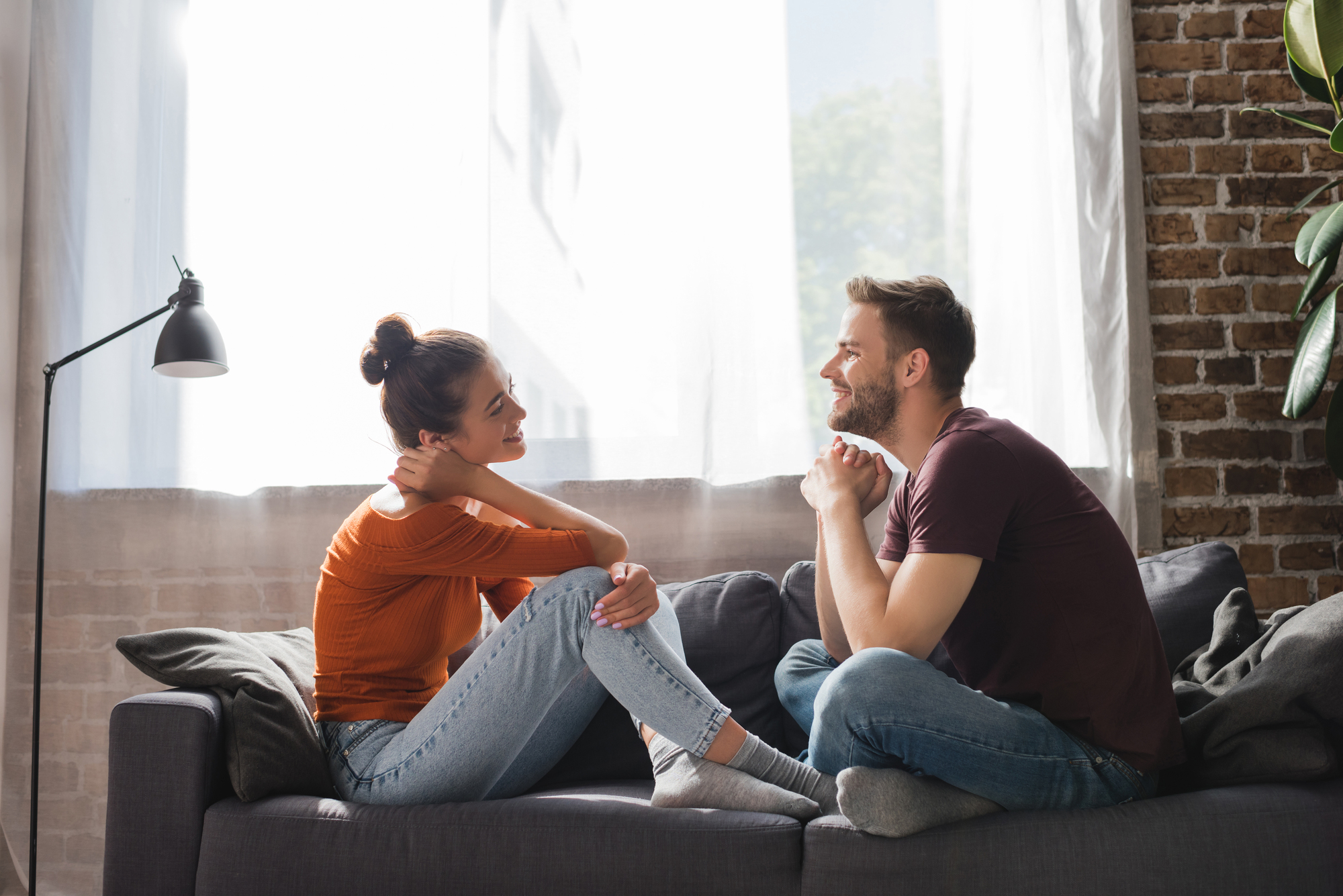 A woman and a man are sitting on a couch facing each other in a cozy, well-lit room with a brick wall and large windows. They both appear to be smiling and engaged in a relaxed conversation. A lamp and some indoor plants are visible in the background.