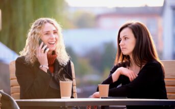 Two women are sitting at an outdoor café with two disposable cups on the table. One woman with curly blonde hair is smiling and talking on a cellphone, while the other woman with straight dark hair looks at her with a neutral expression.