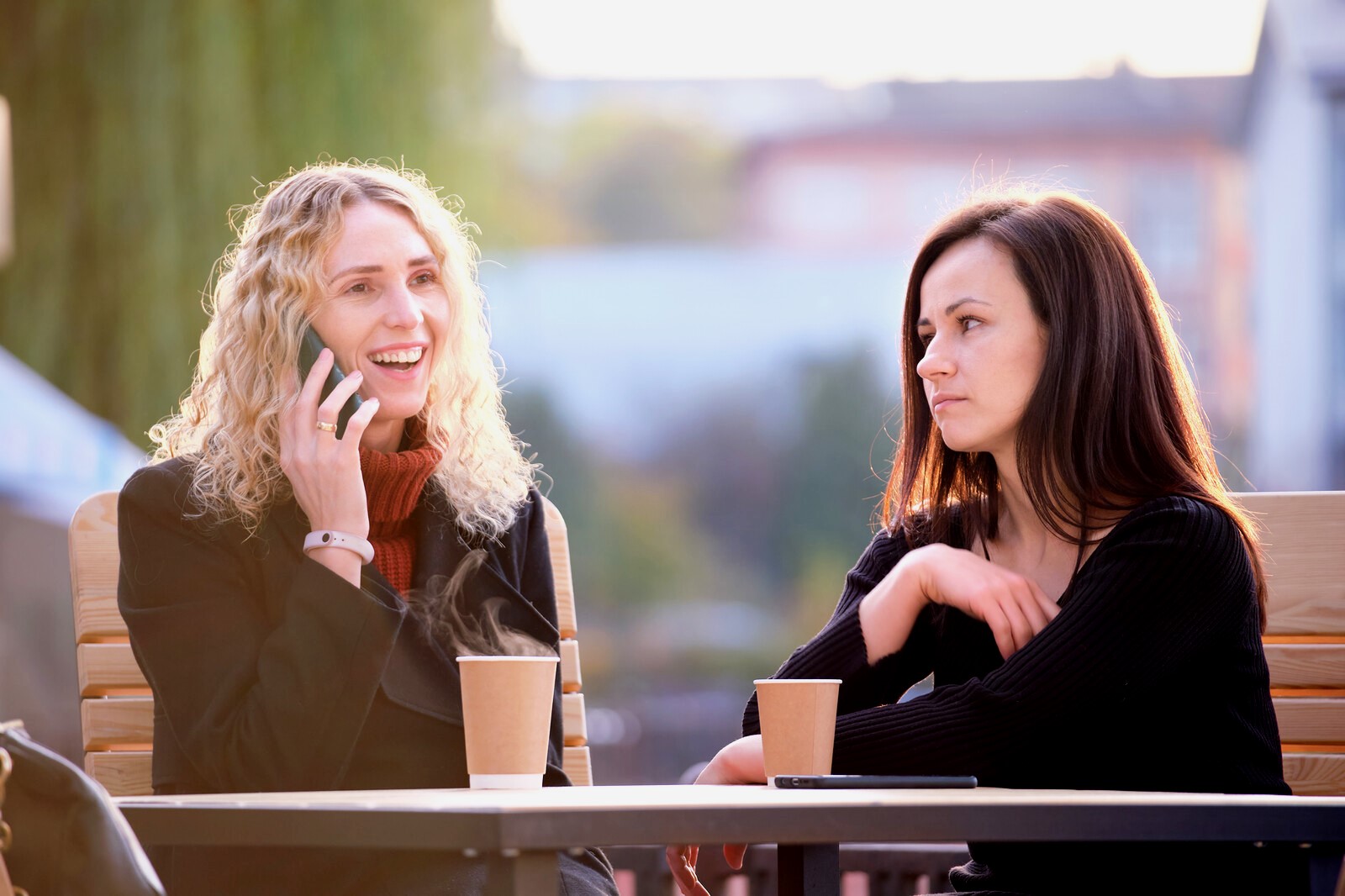 Two women are sitting at an outdoor café with two disposable cups on the table. One woman with curly blonde hair is smiling and talking on a cellphone, while the other woman with straight dark hair looks at her with a neutral expression.