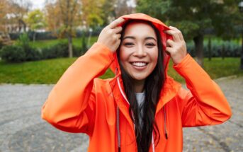 A person smiling and holding the hood of their bright orange rain jacket. They are standing outdoors in a park-like setting with green grass and trees in the background. The weather appears to be overcast.