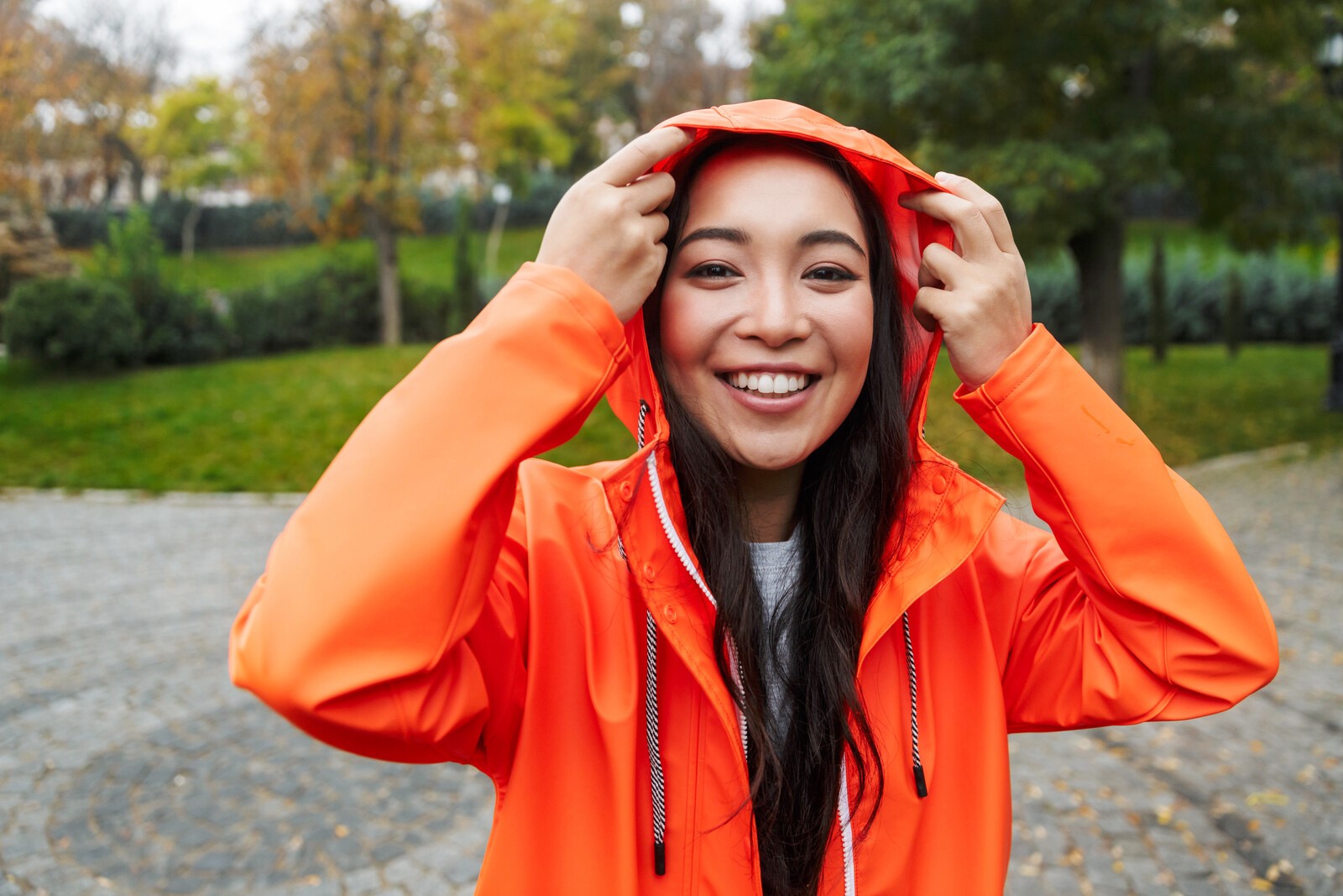 A person smiling and holding the hood of their bright orange rain jacket. They are standing outdoors in a park-like setting with green grass and trees in the background. The weather appears to be overcast.