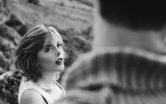 A black-and-white image of a woman with short, tousled hair looking towards the camera. She has a tattoo on her shoulder. Part of a person's back is visible in the foreground, but the focus remains on the woman. The setting appears to be outdoors.