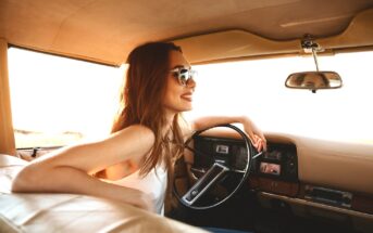 A young woman with long, light brown hair sits in the driver's seat of a vintage car, smiling and wearing sunglasses. Sunlight pours in through the car’s windows, casting a warm glow on her and the car's tan interior.