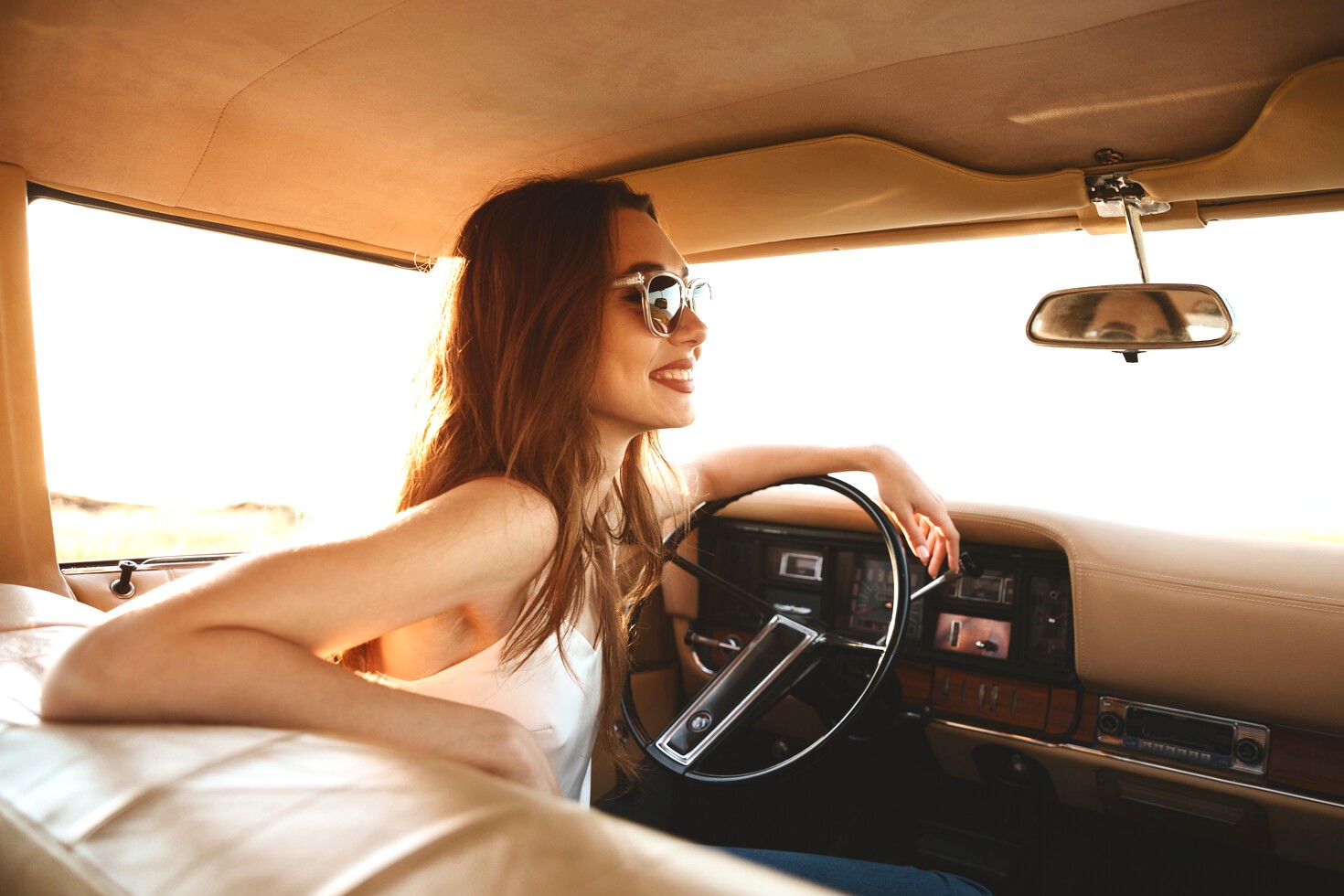 A young woman with long, light brown hair sits in the driver's seat of a vintage car, smiling and wearing sunglasses. Sunlight pours in through the car’s windows, casting a warm glow on her and the car's tan interior.