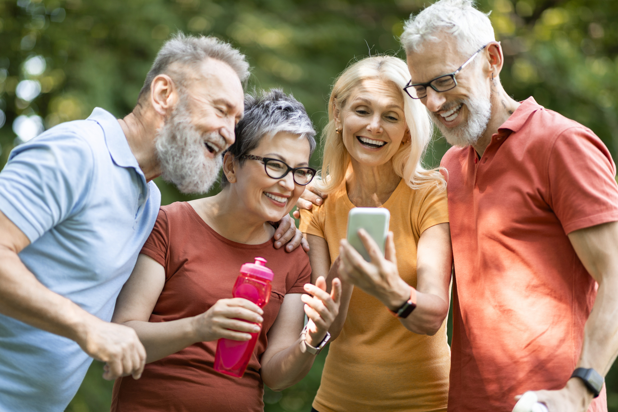 A group of four elderly friends, two men and two women, smile and laugh while looking at a smartphone outdoors. One woman holds a pink water bottle. They are casually dressed and standing on a sunny day with trees in the background.