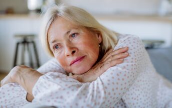 An elderly woman with light-colored hair and blue eyes is resting her head on her folded arms. She wears a white shirt with pink polka dots and has a thoughtful, wistful expression. The background is softly blurred, suggesting an indoor setting.