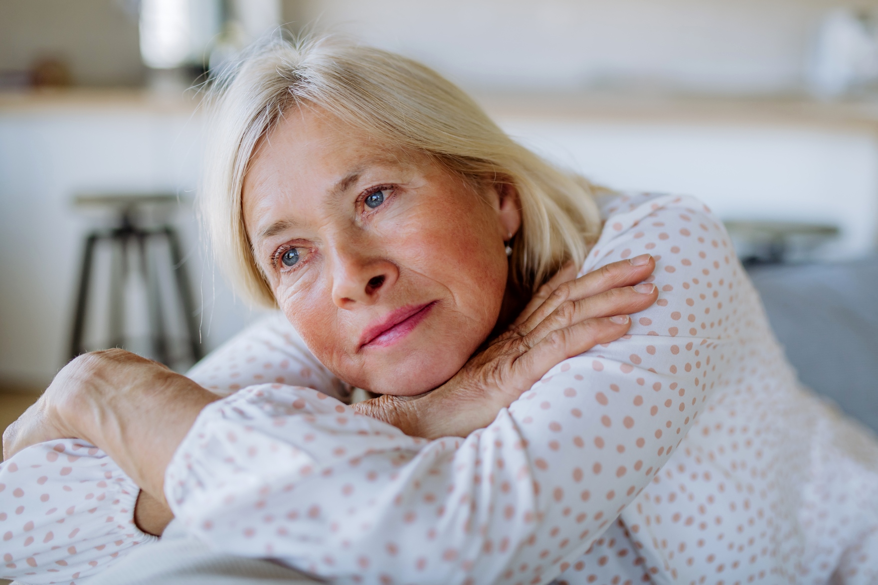 An elderly woman with light-colored hair and blue eyes is resting her head on her folded arms. She wears a white shirt with pink polka dots and has a thoughtful, wistful expression. The background is softly blurred, suggesting an indoor setting.