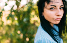 A woman with long dark hair stands outdoors, wearing a blue striped shirt. Her hair is gently blown to the side by the wind. The background is bright and blurred with lush greenery, creating a bokeh effect.