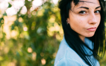 A woman with long dark hair stands outdoors, wearing a blue striped shirt. Her hair is gently blown to the side by the wind. The background is bright and blurred with lush greenery, creating a bokeh effect.