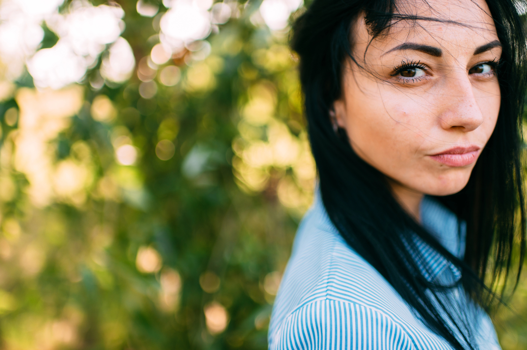 A woman with long dark hair stands outdoors, wearing a blue striped shirt. Her hair is gently blown to the side by the wind. The background is bright and blurred with lush greenery, creating a bokeh effect.