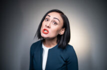 A woman with short black hair, wearing a navy blue jacket and white top, is making an expressive face with raised eyebrows and lips parted as if speaking or singing. She stands against a gray background.