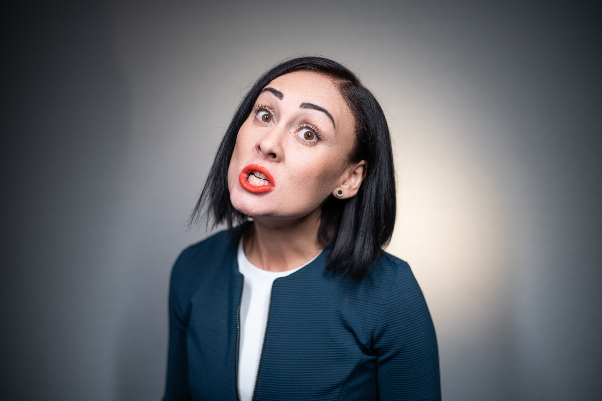 A woman with short black hair, wearing a navy blue jacket and white top, is making an expressive face with raised eyebrows and lips parted as if speaking or singing. She stands against a gray background.