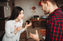 A woman with long dark hair, wearing a white shirt, is standing in a kitchen with her hands raised in an expressive manner, speaking to a man with short hair in a plaid shirt. They appear to be in a serious conversation. The background shows a table and chairs.