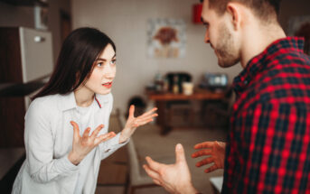 A woman with long dark hair, wearing a white shirt, is standing in a kitchen with her hands raised in an expressive manner, speaking to a man with short hair in a plaid shirt. They appear to be in a serious conversation. The background shows a table and chairs.