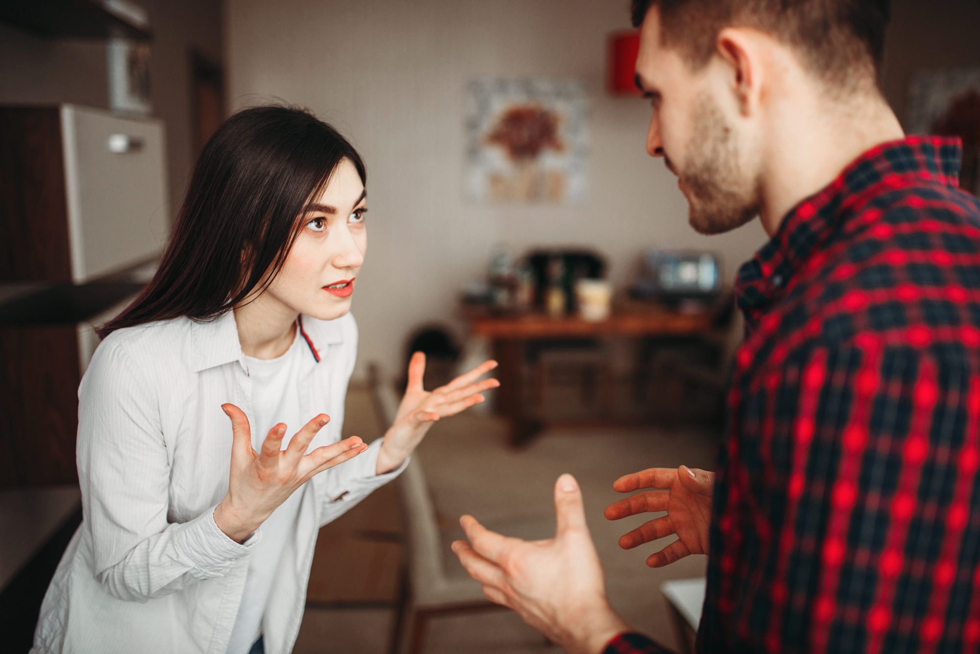 A woman with long dark hair, wearing a white shirt, is standing in a kitchen with her hands raised in an expressive manner, speaking to a man with short hair in a plaid shirt. They appear to be in a serious conversation. The background shows a table and chairs.