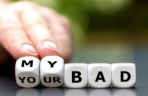 A hand is pushing aside lettered dice blocks arranged to spell "MY YOUR BAD" with the emphasis on "MY BAD." The background is blurred with a mix of green and white, suggesting an outdoor setting.