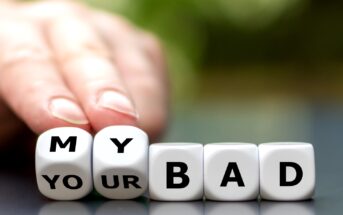 A hand is pushing aside lettered dice blocks arranged to spell "MY YOUR BAD" with the emphasis on "MY BAD." The background is blurred with a mix of green and white, suggesting an outdoor setting.