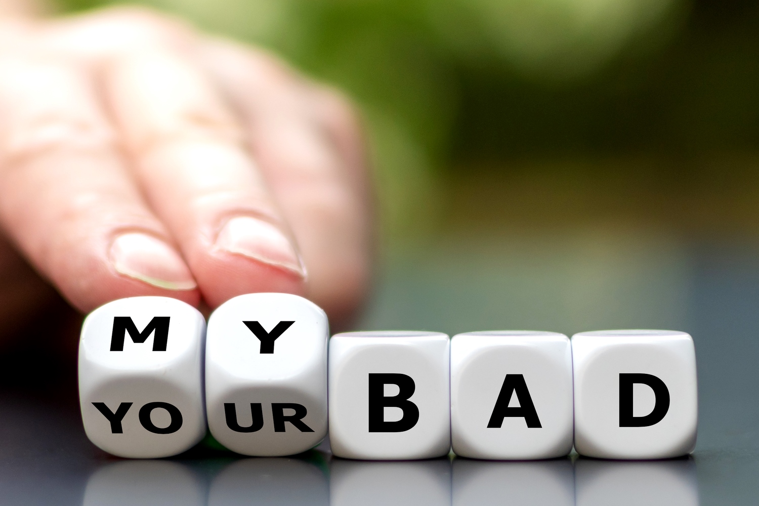 A hand is pushing aside lettered dice blocks arranged to spell "MY YOUR BAD" with the emphasis on "MY BAD." The background is blurred with a mix of green and white, suggesting an outdoor setting.